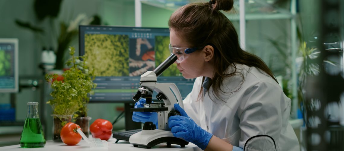 Portrait of biologist scientist in white coat working in expertise laboratory looking into microscope analyzing organic gmo leaf. Specialist researcher doing biochemistry experiment